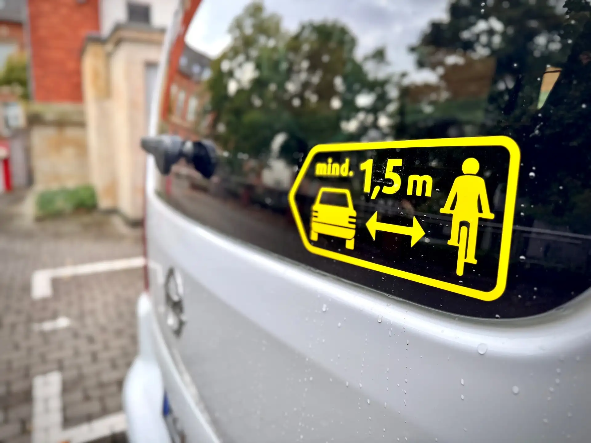 Gelb-schwarzer Aufkleber mit Auto-, Fahrrad- und Doppelpfeil-Symbol sowie Text „Mind. 1,5 m“.