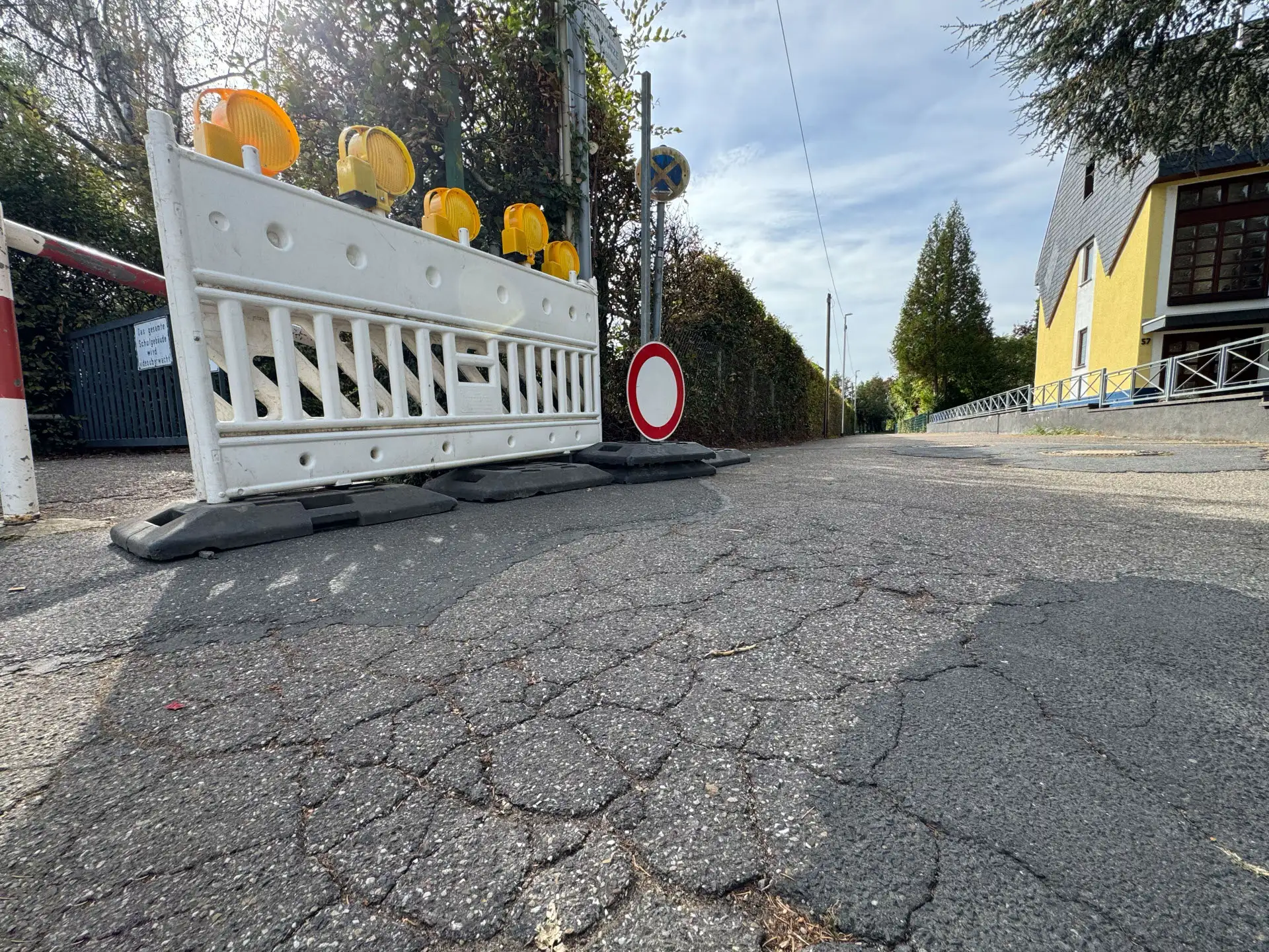 Eine weiße Baustellenabsperrung mit gelben Warnleuchten steht auf rissigem Asphalt vor der Zufahrt zur Grundschule Kesselheim.