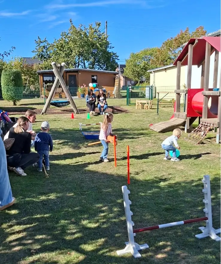 Kinder spielen auf der Wiese im Außengelände der KiTa an einem Parcours mit Stangen und Hürden; Erwachsene schauen zu.