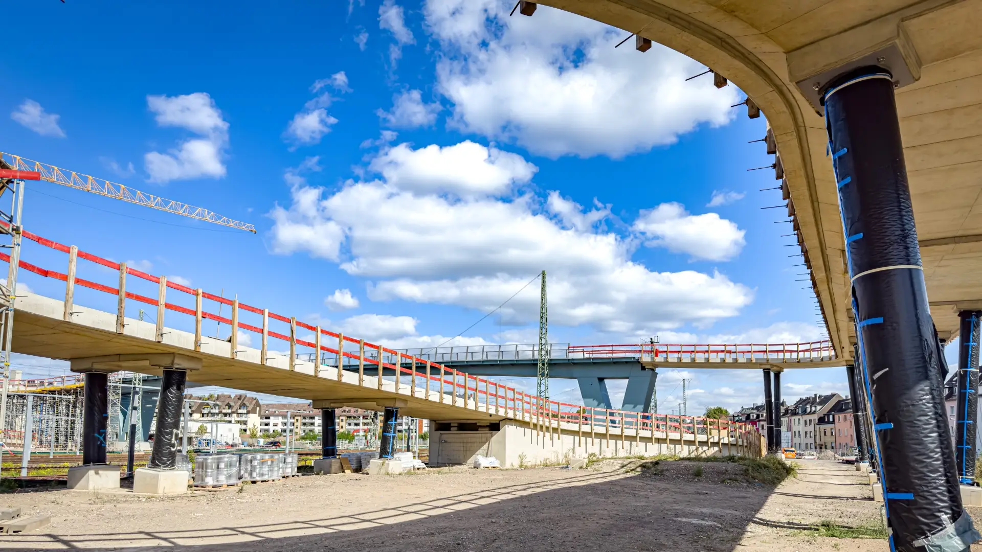 Die im Bau befindliche Geh- und Radwegbrücke mit Betonrampen und roten Schutzgeländern überspannt ein Bahngelände.
